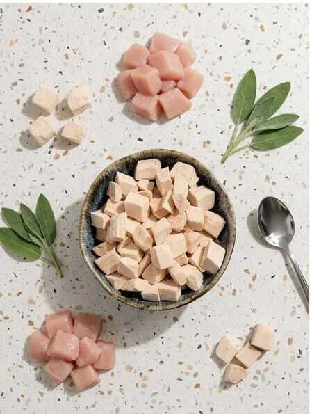 PawDry Freeze-Dried Chicken Bites Pets Treat 100g in a bowl surrounded by fresh leaves and bite-sized pieces on a white surface.