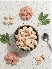PawDry Freeze-Dried Chicken Bites Pets Treat 100g in a bowl surrounded by fresh leaves and bite-sized pieces on a white surface.