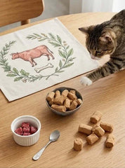 Cat reaching for snacks on a table with PawDry Freeze-Dried Beef Raw Meaty Bone (RMB) 100g.