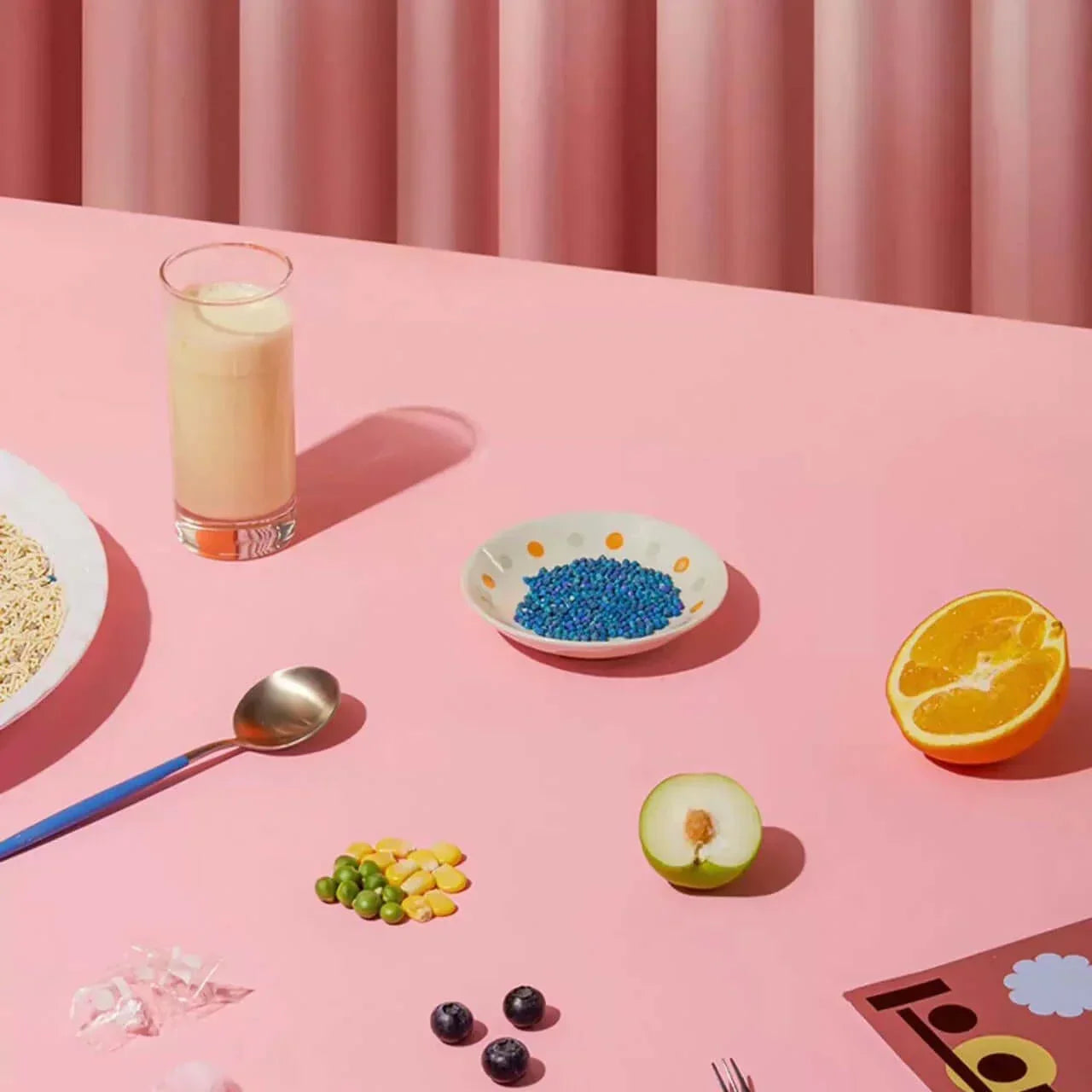 Pink table with colorful food items: blue beads, orange, apple, berries, peas, corn, milk, and noodles
