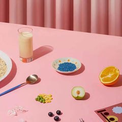 Pink table with colorful food items: blue beads, orange, apple, berries, peas, corn, milk, and noodles