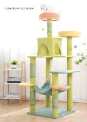 A multi-level cat tree with a light green base and posts, pastel yellow and peach platforms, a mint green hammock, and a small white cat sitting on the top perch. The scene is set in a bright, modern room with a wooden floor, a small wooden shelf with books and a plant, and white curtains in the background.