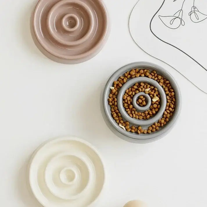 Spiral slow feeder dog bowls in beige, gray, and cream with kibble, styled on white surface