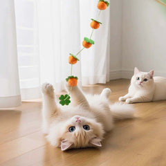Fluffy cat playing with orange pom-pom cat toy on wooden floor, another cat watching nearby