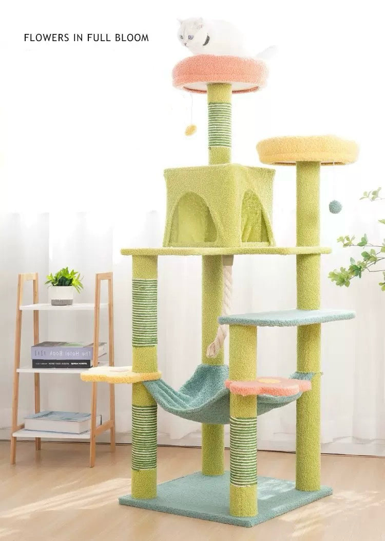 A multi-level cat tree with a light green base and posts, pastel yellow and peach platforms, a mint green hammock, and a small white cat sitting on the top perch. The scene is set in a bright, modern room with a wooden floor, a small wooden shelf with books and a plant, and white curtains in the background.