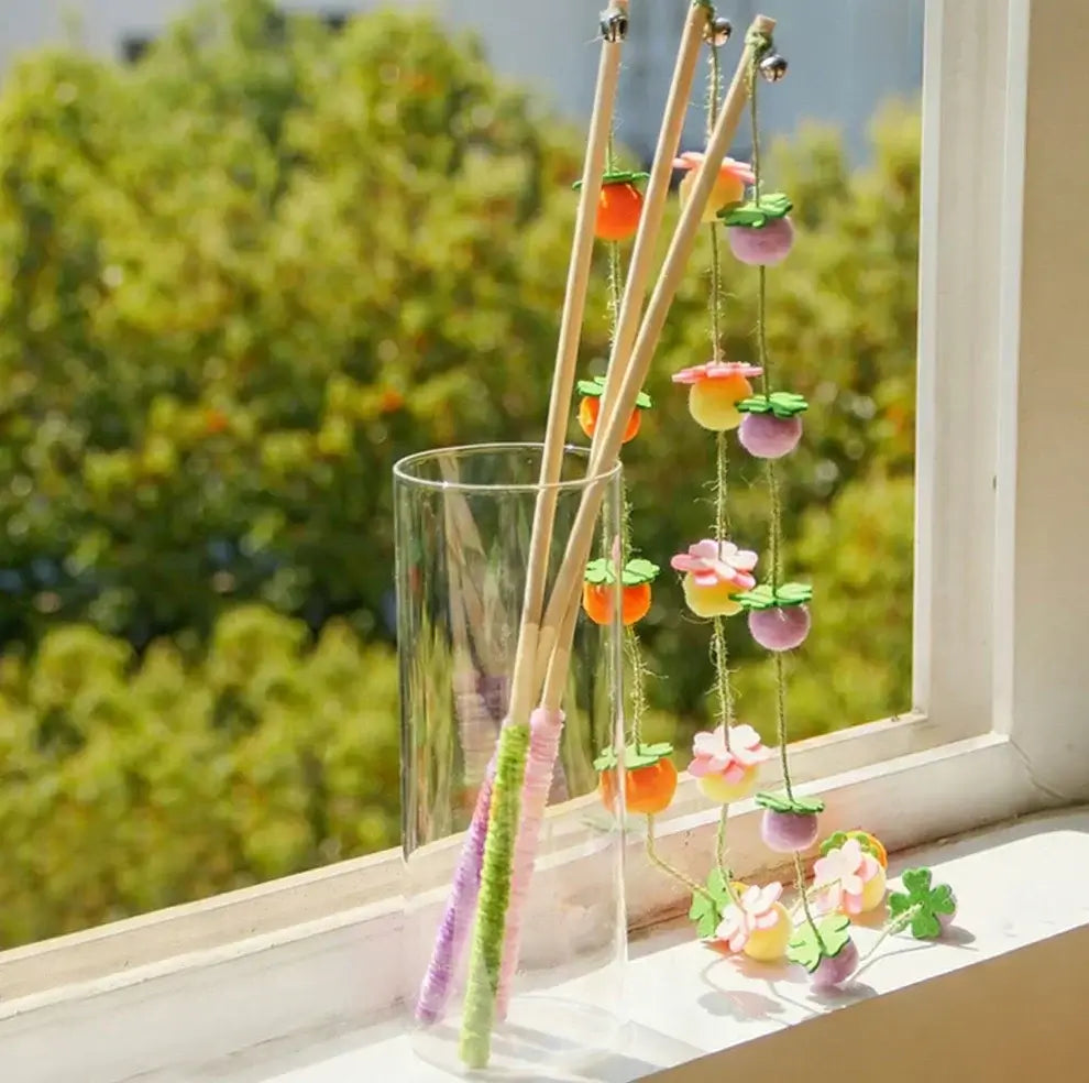 Colorful cat wand toys with felt flowers and bells, displayed in a glass vase by a sunny window.